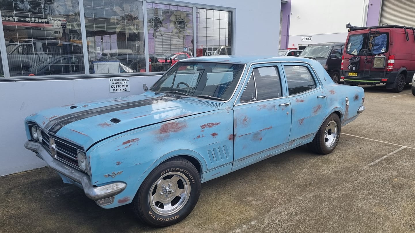 Light blue Holden HZ Premier with patina parked in front of the R & M shopfront